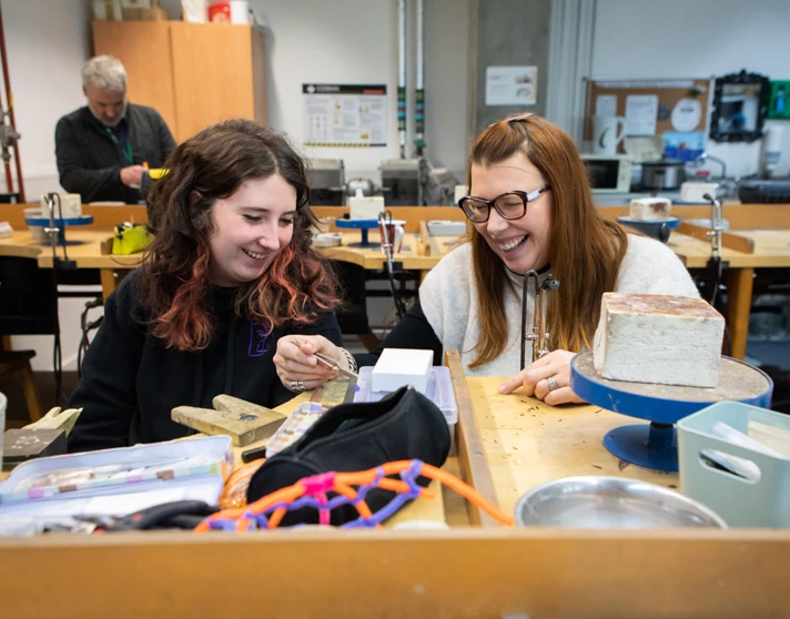 Two women in a jewellery workshop laughing and working together at a wooden workbench, with various tools and equipment scattered around. Two women in a jewellery workshop laughing and working together at a wooden workbench, with various tools and equipment scattered around.